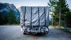 Deux vélos électriques recouverts d’une housse grise renforcée et solidement sanglée sur le porte-vélos arrière d’un camping-car, dans une aire de stationnement de montagne.
